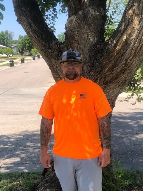 A man in an orange shirt standing next to a tree.